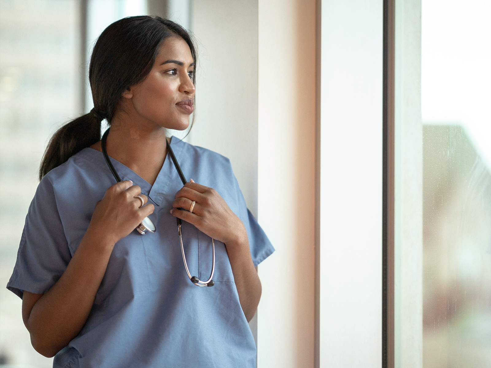Kvinnlig läkare i blå läkartröja och stetoskop runt halsen blickar ut genom ett fönster