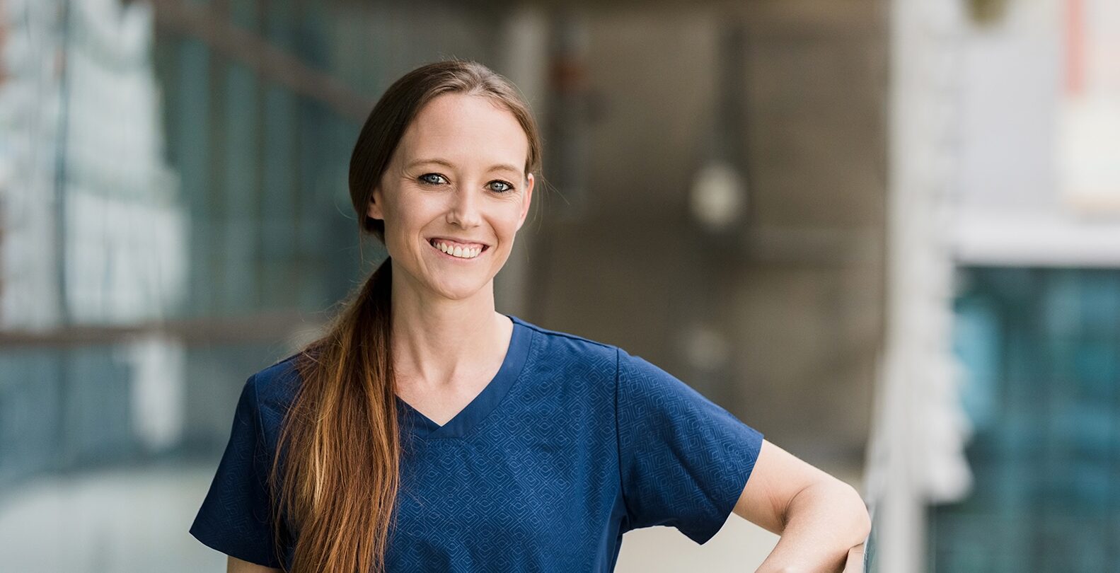 Kvinnlig läkare med blå tröja och långt hår i tofs ler mot kameran